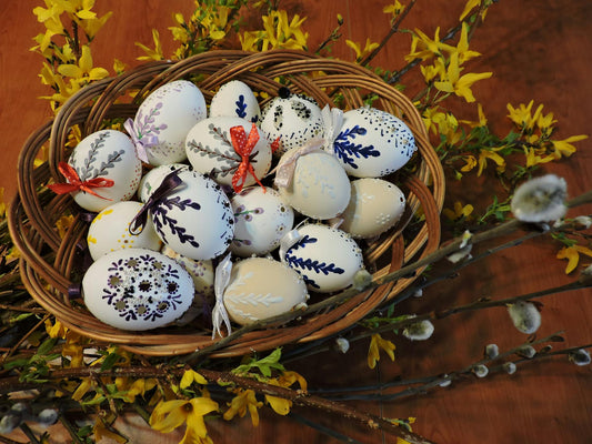 Wicker basket filled with hand-painted Easter eggs in cream and white, decorated with navy and lavender botanical patterns, dotted details, and satin ribbon bows; warm indoor light on wood backdrop with bright yellow spring blossoms