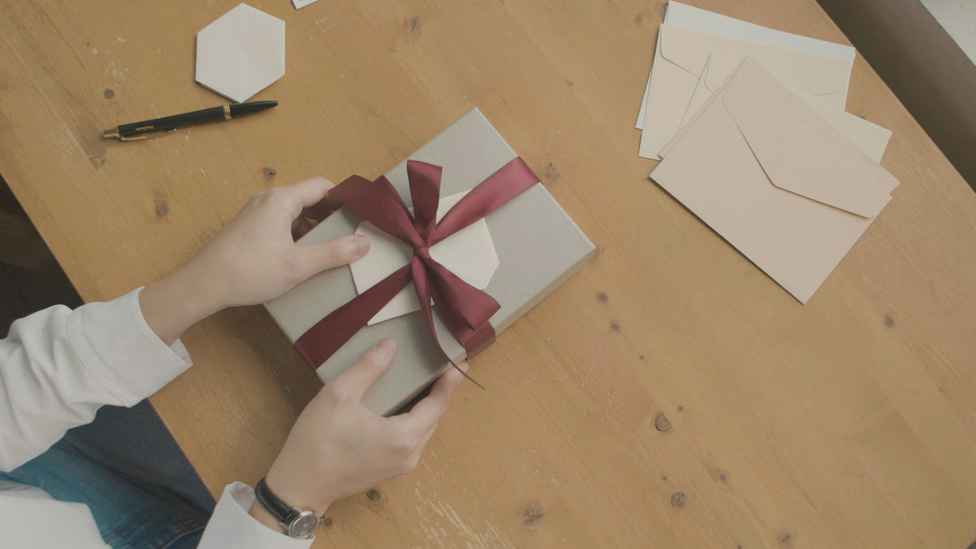 Hands holding a neatly wrapped gift box with a red ribbon and blank card on a wooden table.