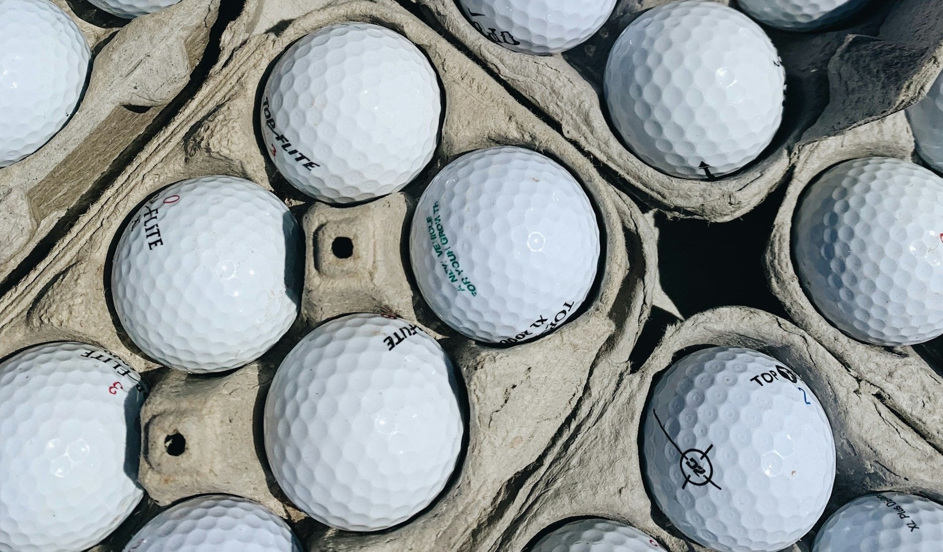 Close view of white golf balls imprinted with various black and green text, neatly arranged in a carton-style holder for transportation or storage.
